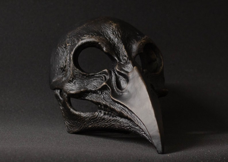 May include: A dark, textured mask with a bird-like beak and eye openings. The mask is a dark, matte color, possibly bronze or black, and has a detailed, weathered appearance, suggesting a gothic or historical theme.