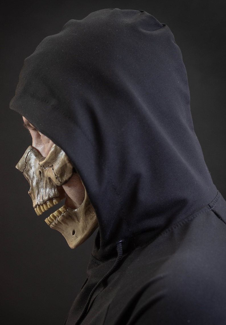 Pu&ograve; includere: Un profilo laterale di una persona che indossa una felpa con cappuccio nera e una maschera a teschio. La maschera &egrave; una replica dettagliata di un teschio umano, con denti e struttura ossea visibili. Lo sfondo &egrave; grigio scuro.
