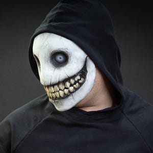 May include: A white mask with a sinister grin and large teeth, worn with a black hooded sweatshirt. The mask has a cracked texture and blank, pale eyes, creating a spooky, Halloween-themed appearance.