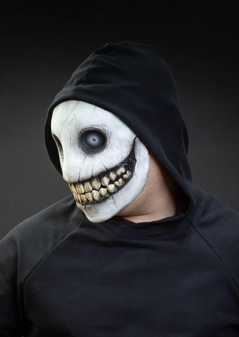 May include: A white mask with a sinister grin and large teeth, worn with a black hooded sweatshirt. The mask has a cracked texture and blank, pale eyes, creating a spooky, Halloween-themed appearance.