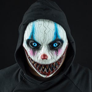 Peut inclure: Un masque de clown blanc à la texture craquelée, aux yeux bleu vif lumineux, aux orbites sombres, au nez rouge et au large sourire denté aux dents pointues tachées de sang. Le masque est porté avec un vêtement noir à capuche couvrant la tête.
