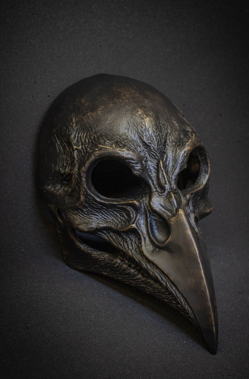 May include: A dark bronze-colored mask with a bird-like beak and eye sockets. The mask has a textured surface and is likely made of metal or a similar material. The mask is designed to cover the face.