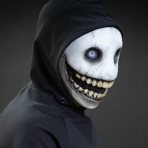 May include: A white mask with a wide, unsettling grin and large, vacant eyes. The mask is worn with a black hooded sweatshirt. The mask has a cracked texture and detailed teeth, creating a spooky appearance.