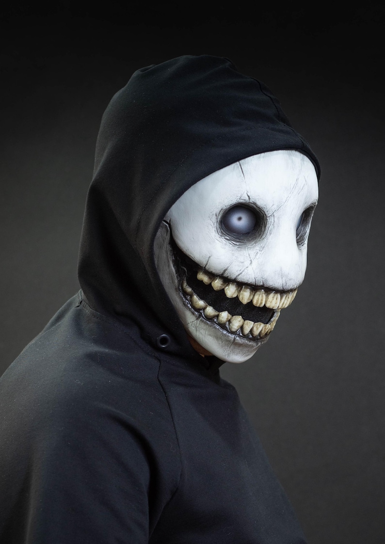 May include: A white mask with a wide, unsettling grin and large, vacant eyes. The mask is worn with a black hooded sweatshirt. The mask has a cracked texture and detailed teeth, creating a spooky appearance.