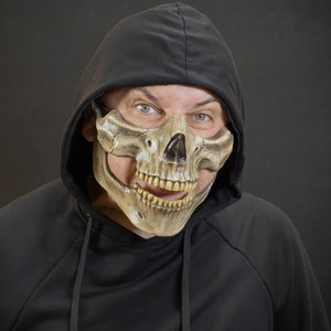 Pu&ograve; includere: Una maschera a teschio color cuoio con naso e denti neri &egrave; indossata sulla parte inferiore del viso. La maschera &egrave; indossata con una felpa con cappuccio nera. Lo sfondo &egrave; grigio scuro.