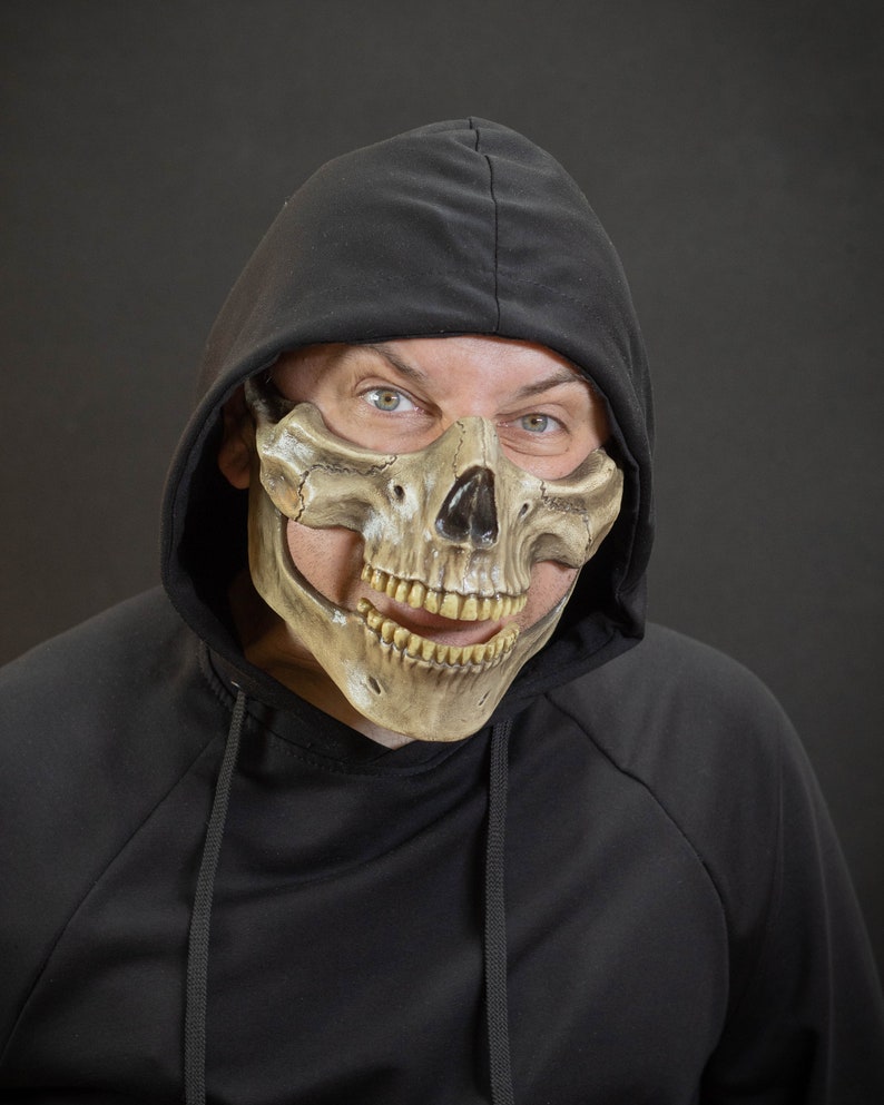 Pu&ograve; includere: Una maschera a teschio color cuoio con naso e denti neri &egrave; indossata sulla parte inferiore del viso. La maschera &egrave; indossata con una felpa con cappuccio nera. Lo sfondo &egrave; grigio scuro.