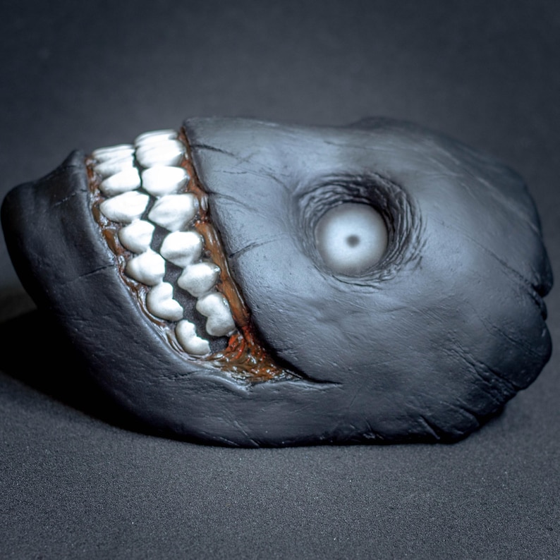 May include: A black and gray mask with a large, toothy grin and a single white eye. The mask is made of a textured material and has a slightly grotesque appearance.