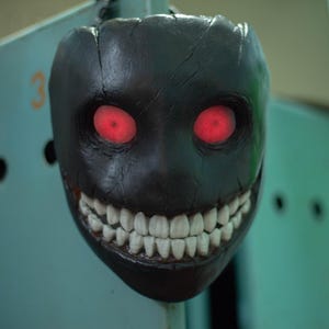 May include: A black mask with a wide, toothy grin and glowing red eyes. The mask is made of a textured material and has a sinister appearance.