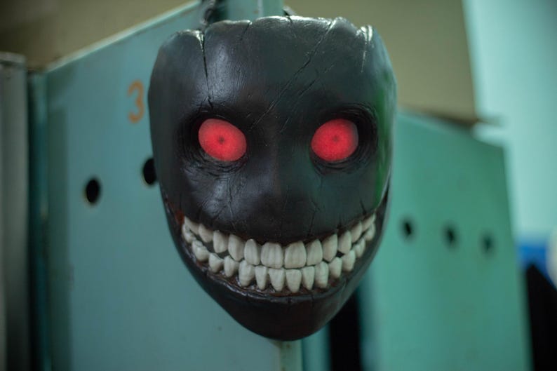 May include: A black mask with a wide, toothy grin and glowing red eyes. The mask is made of a textured material and has a sinister appearance.