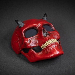 May include: A red devil skull mask with black horns and white teeth. The mask is made of a hard plastic material and has a glossy finish.