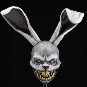 Op de afbeelding: Een konijnenmasker voor het hele hoofd met lange, rechtopstaande witte vachtoren en zwarte binnenkanten. Het grijze gezicht heeft een dreigende uitdrukking, wit oplichtende ogen, een roze neus en een mond vol scherpe, gele, grillige tanden.