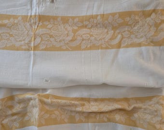 May include: A white textile fabric featuring horizontal bands of soft yellow. The yellow sections are decorated with a woven floral pattern depicting roses and leaves. The fabric shows visible folds and slight texture throughout the surface.