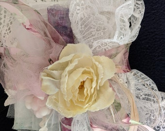 May include: A close-up of a decorative floral arrangement featuring a large, pale yellow rose as the focal point. The arrangement includes layers of white lace, pink tulle, and ribbons in various shades, with a touch of purple mesh.