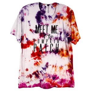 May include: A white t-shirt with a tie-dye pattern in shades of red, orange, purple, and pink. The text "MEET ME at the PUMPKIN PATCH" is printed in black across the front of the shirt.