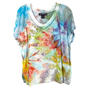 May include: A tie-dye V-neck t-shirt with short sleeves. The shirt features a colorful pattern with shades of blue, red, yellow, and green. A white embroidered design is on the front. The shirt has a white trim around the neckline.