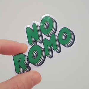 May include: A sticker with the words "NO ROMO" in green, outlined in white and black, with a 3D effect. The sticker is held between fingers against a white background.