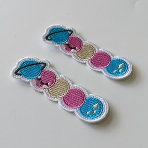 May include: Two embroidered patches featuring a line of planets in blue, pink and beige. Each patch has a planet with a ring around it at the start of the line.