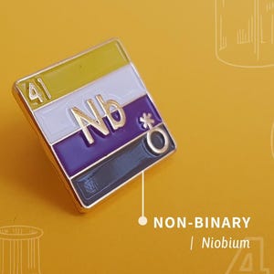 May include: A gold-coloured pin with a yellow, white, purple, and black striped design. The pin has the chemical symbol "Nb" and the number "41" on it. The text "NON-BINARY | Niobium 41" is below the pin.