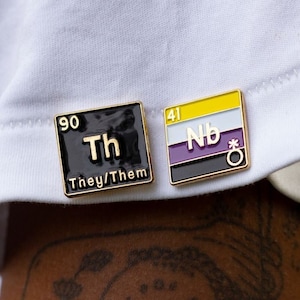 Peut inclure: Deux épingles dorées sur une chemise blanche. L'épingle de gauche est noire avec le texte "90 Th They/Them". L'épingle de droite est jaune, blanche et violette avec le texte "41 Nb" et un symbole.