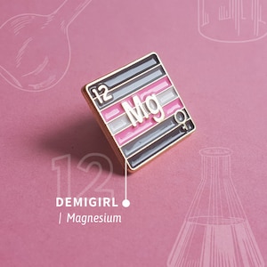 Peut inclure: Une épingle en émail de forme carrée de couleur dorée. L'épingle présente un fond à rayures noires et blanches avec une bande rose au milieu. Les lettres "Mg" sont au centre de l'épingle, avec le numéro "12" en haut à gauche. Le texte "DEMIGIRL | Magnésium" est sous l'épingle.