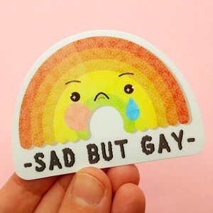 May include: A rainbow sticker with a sad cartoon face and the text "Sad but Gay" in black.