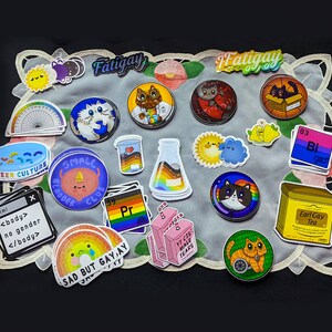 May include: A collection of colourful stickers featuring various designs, including cats, a rainbow, a beaker, a sun, a cloud, and text. The stickers are arranged on a white doily with a black background.