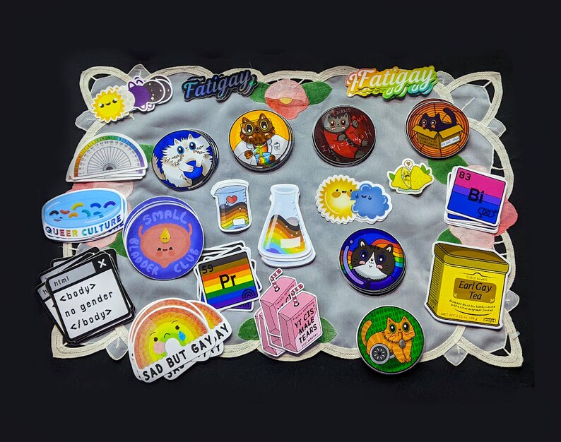 May include: A collection of colourful stickers featuring various designs, including cats, a rainbow, a beaker, a sun, a cloud, and text. The stickers are arranged on a white doily with a black background.