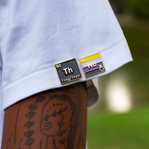 May include: Two enamel pins in the shape of periodic table elements. The first pin is black with gold trim and says "Th" with the number 90 above it and "They/Them" below it. The second pin is yellow with black and purple stripes and says "Nb" with the number 41 above it and a gender neutral symbol below it.