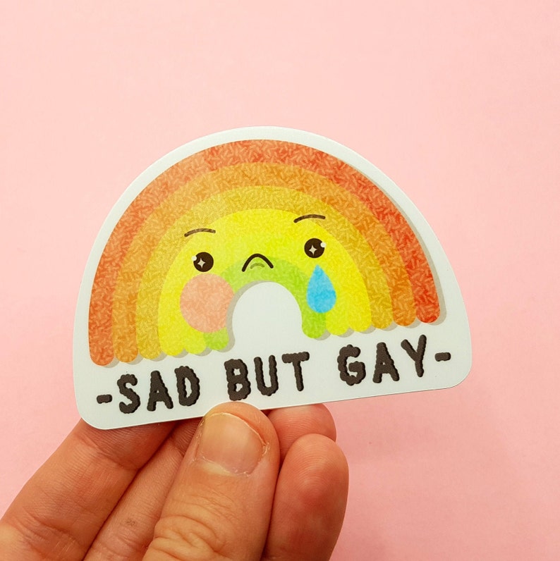 May include: A rainbow sticker with a sad face and a tear. The text on the sticker says "Sad But Gay".