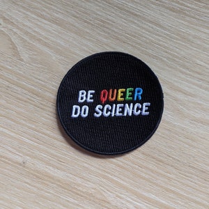 May include: Black round patch with white embroidered text that reads "BE QUEER DO SCIENCE". The word "QUEER" is embroidered in rainbow colours.