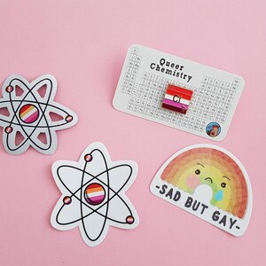 May include: Three stickers featuring a rainbow, an atom with a lesbian pride flag, and an atom with a bisexual pride flag. The rainbow sticker has a cartoon face with a sad expression and the text "Sad But Gay". The periodic table sticker has a lesbian pride flag in the box for chlorine.