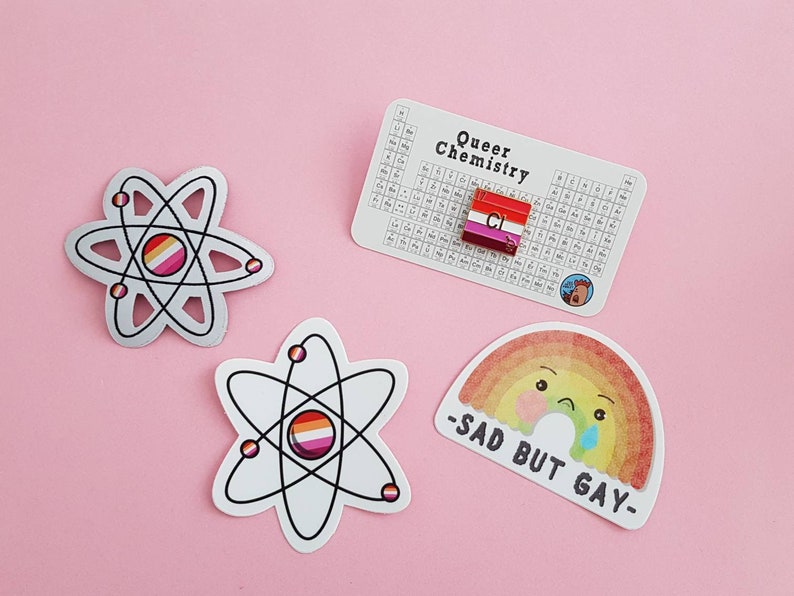 May include: Three stickers featuring a rainbow, an atom with a lesbian pride flag, and an atom with a bisexual pride flag. The rainbow sticker has a cartoon face with a sad expression and the text "Sad But Gay". The periodic table sticker has a lesbian pride flag in the box for chlorine.