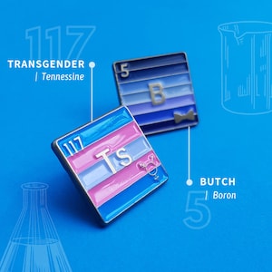 Op de afbeelding: Twee emaillepins met een wetenschappelijk ontwerp. De eerste pin is blauw en paars met de letter "B" en het nummer "5" en de tekst "BUTCH" en "Boron". De tweede pin is roze, blauw en wit met de letters "Ts" en het nummer "117" en de tekst "TRANSGENDER" en "Tennesseine".