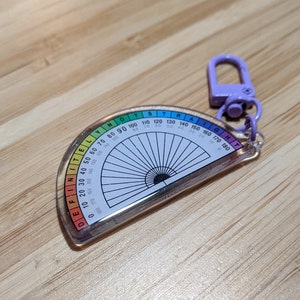 Peut inclure: Un porte-clés en plastique transparent en forme de rapporteur d'angles avec un bord arc-en-ciel et des paillettes incrustées dans le plastique. Le rapporteur mesure les angles de 0 à 180 degrés et porte l'inscription "DEFINITELY NOT STRAIGHT" sur le bord.