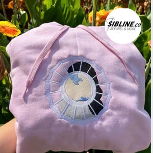 K&ouml;nnte beinhalten: Rosafarbener Hoodie mit einem wei&szlig;en und blauen gestickten Design eines Gesichts in einem Kreis. Der Text "SIBLINE.CO APPAREL & MORE" befindet sich in der oberen rechten Ecke des Bildes.