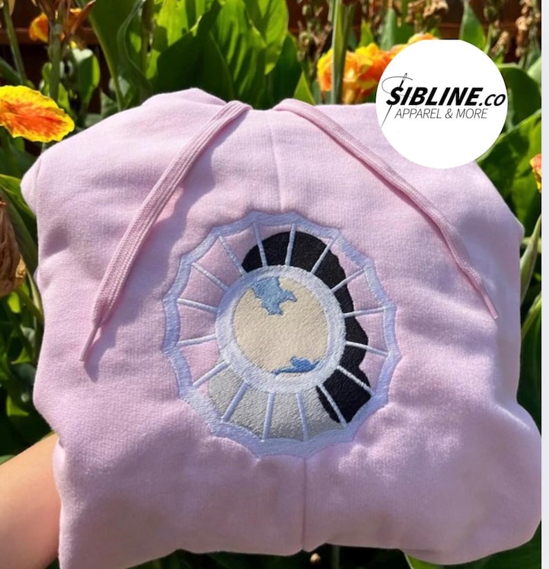 K&ouml;nnte beinhalten: Rosafarbener Hoodie mit einem wei&szlig;en und blauen gestickten Design eines Gesichts in einem Kreis. Der Text "SIBLINE.CO APPAREL & MORE" befindet sich in der oberen rechten Ecke des Bildes.