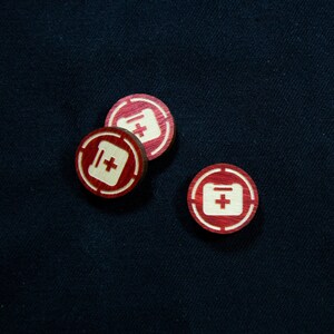May include: Three red wooden tokens with a white plus sign inside a white rectangle. The tokens are on a dark blue surface.