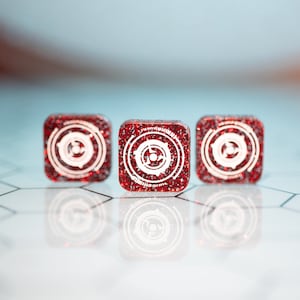 May include: Three red square-shaped dice with white circular designs on each face. The dice are on a white surface with a hexagonal pattern.