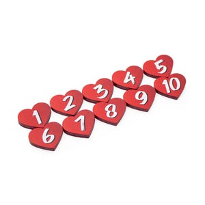 May include: A collection of ten red heart-shaped wooden pieces, each featuring a white numeral from one to ten. The hearts are arranged in a scattered formation, displaying the numbers clearly against the solid red background of each heart shape.