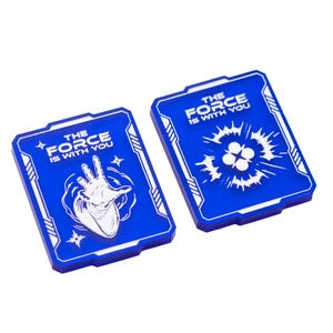 May include: Two blue rectangular tokens with white designs and text. The text on each token reads "THE FORCE IS WITH YOU." One token features a hand gesture, the other a four-petal design. The tokens are likely collectibles or game pieces.