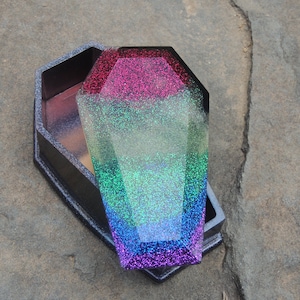 May include: A black coffin-shaped box with a glittery rainbow lid. The lid is open and the inside of the box is black.
