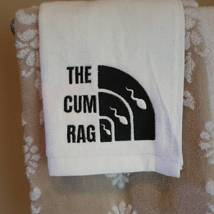 May include: White towel with the text "THE CUM RAG" in black block letters. The design includes a black semi-circle with three sperm cell illustrations. The towel is hanging on a silver bar with a decorative towel in the background.
