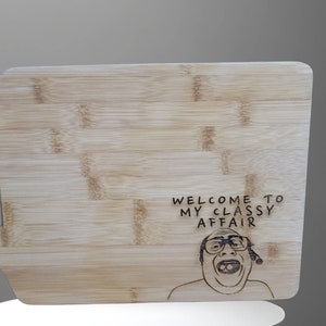 Puede incluir: Una tabla de cortar de madera con un asa. La tabla tiene una imagen grabada con láser de un hombre con el texto "Welcome to my classy affair".