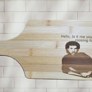 Puede incluir: Una tabla de cortar de madera con mango, que presenta una imagen grabada con láser de un hombre con el texto "Hello, Is it me you're cooking for?"