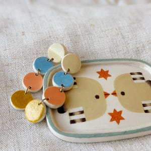 Puede incluir: Pendientes coloridos con colgantes circulares en azul, naranja y dorado. Junto a ellos, una pequeña bandeja rectangular de cerámica con una ilustración de pájaros y estrellas naranjas. La bandeja tiene un borde verde claro.