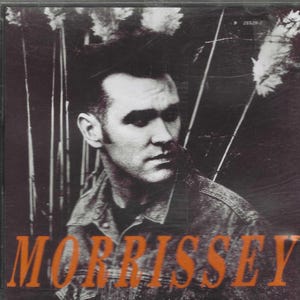 May include: CD case featuring a black and white portrait of a man with dark hair and a denim jacket. The name "MORRISSEY" is printed in large orange letters across the bottom of the case. The background has a dark, textured appearance.