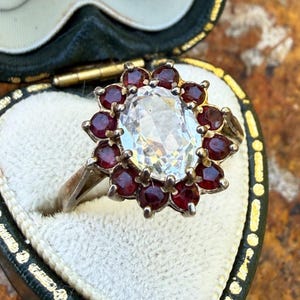 May include: A silver-toned ring featuring a large, oval-cut clear centre stone surrounded by a halo of twelve small, round-cut deep red stones. The ring is set in a heart-shaped jewellery box with a white velvet interior and a dark green exterior.