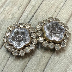 May include: Two vintage flower-shaped buttons. Each button features a clear, faceted center surrounded by a ring of smaller, clear rhinestones. The buttons have a gold-toned metal base and are set against a neutral background.