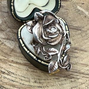 May include: A silver-toned metal brooch shaped like a blooming rose with detailed petals and leaves on a stem. The brooch is resting inside an open, heart-shaped jewellery box that features a dark exterior and a cream-coloured interior lining.