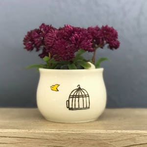 May include: A white ceramic planter with a bird and a birdcage design. The planter is filled with purple flowers.